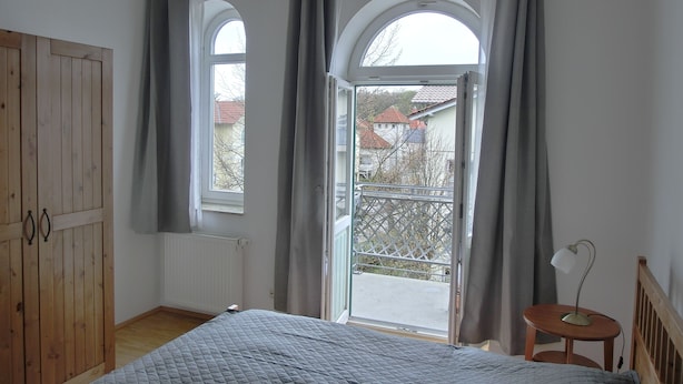 Un dormitorio con cama, armario de madera, ventana con cortinas y balcón con barandilla.