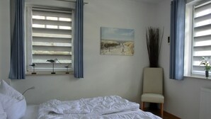 A bedroom with a bed, a chair, a window with blinds, a painting, and a vase with plants. - Zentrale 2 Zimmer-fewo für 4 Pers., Terrasse mit Strandkorb (Graal-Müritz)