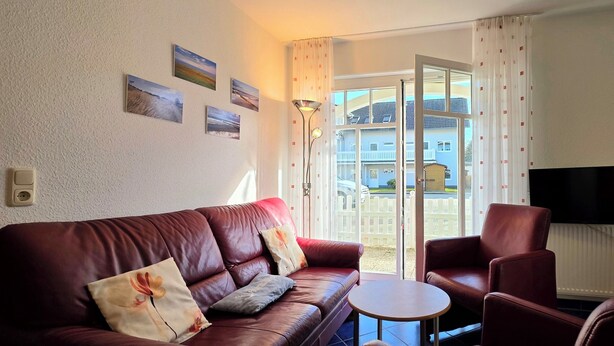 Una sala de estar con un sofá de cuero, una mesita redonda y un sillón de cuero. Hay un televisor montado en la pared y una ventana con cortinas.