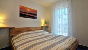Un dormitorio con cabecera de madera, cama con ropa de cama a rayas, dos lámparas de noche, una ventana con cortinas traslúcidas y un cuadro de un atardecer en la pared.