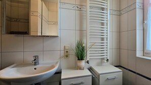 Un baño con un lavamanos blanco, un espejo, un perchero para toallas y una ventana con cortina.