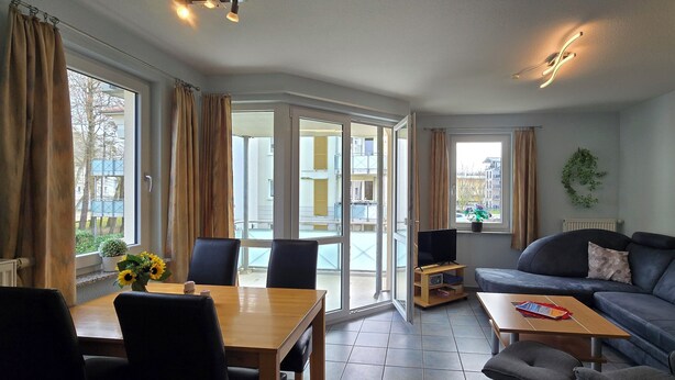 Un salon moderne avec un canapé, une table basse et une table à manger avec des chaises. On y trouve un téléviseur posé sur un meuble et une plante sur le rebord de la fenêtre.