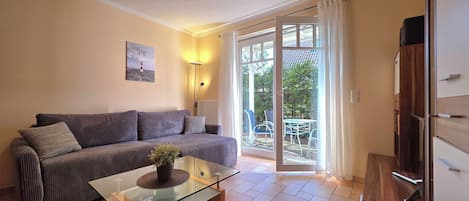A modern living room with a sofa, a glass coffee table with a plant, a lamp, and a sliding glass door leading to an outdoor area with a table and chairs.
