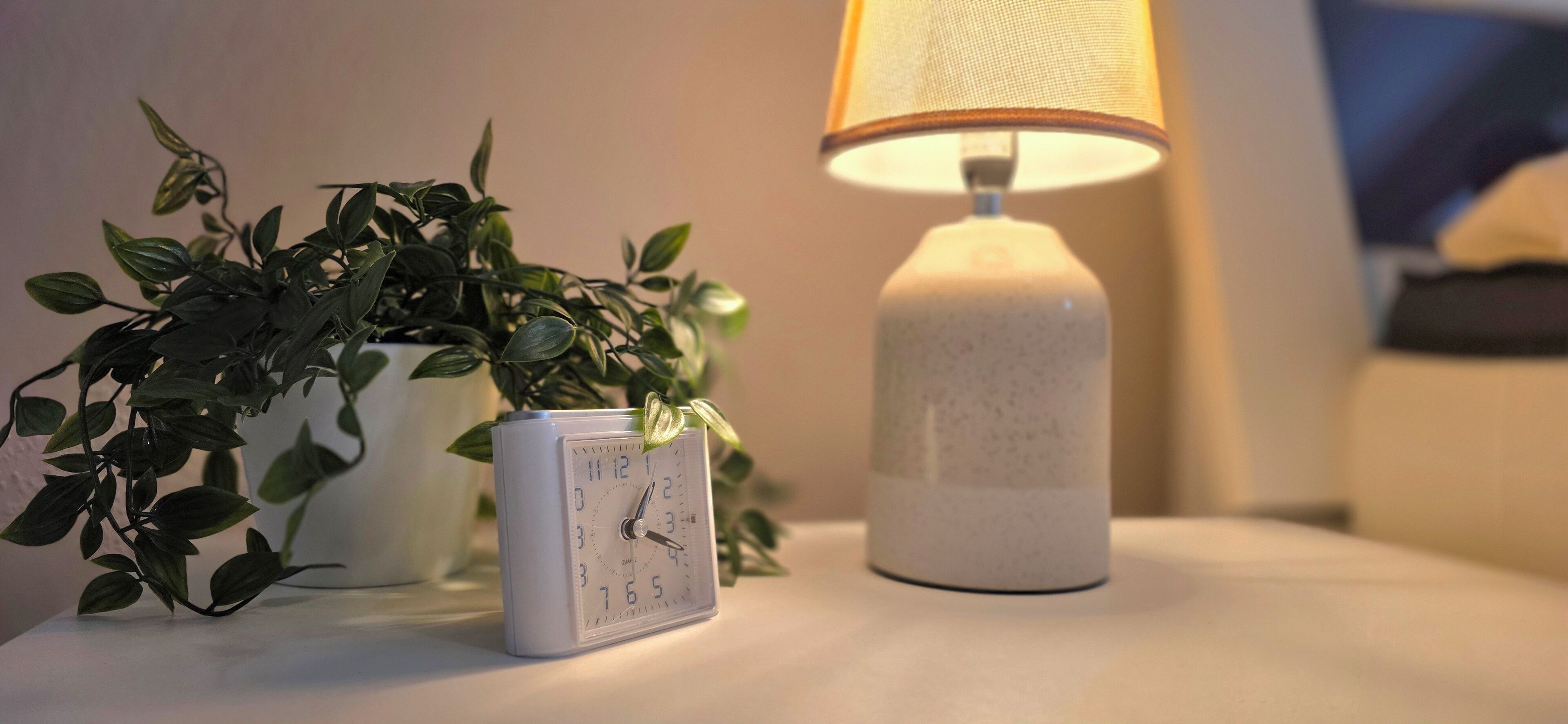 Ein Nachttisch mit einer weißen Uhr, einer weißen Lampe und einer Topfpflanze.