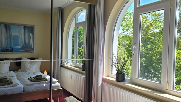 A hotel room with a bed, a painting, a TV, and a view of trees through large windows. - Luxuriöse 3-zi.-fewo für 4 Pers., Inklusive Bettwäsche, Handtücher und Wlan (Graal-Müritz)