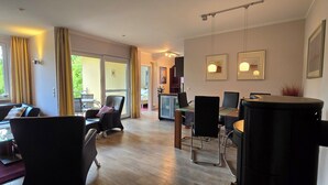 A modern living room with a sofa, chairs, a dining table, and a kitchen area visible through an open door. - Luxuriöse 3-zi.-fewo für 4 Pers., Inklusive Bettwäsche, Handtücher und Wlan (Graal-Müritz)