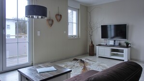 A living room with a sofa, a coffee table, a TV on a stand, a vase with branches, and a sliding glass door leading to an outdoor area.