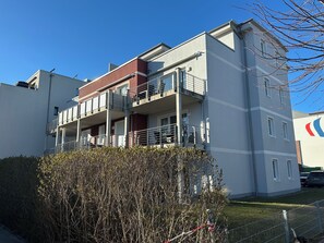 A multi-story residential building with balconies, a clear sky, and a hedge in the foreground.