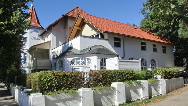 A two-story building with a red-tiled roof, white walls, and a small tower-like structure on the left.