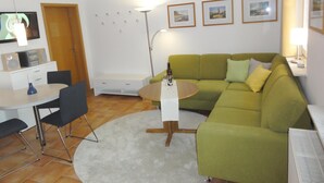 A living room with a green sofa, a white coffee table, a dining table with chairs, a TV, and a lamp.