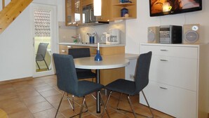 A modern kitchen with wooden cabinets, a dining table with chairs, a TV mounted on the wall, and a built-in speaker.
