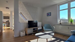 Ein modernes Wohnzimmer mit einem Flachbildfernseher, einem blauen Sessel, einem weißen Sofa und einem Essbereich im Hintergrund.