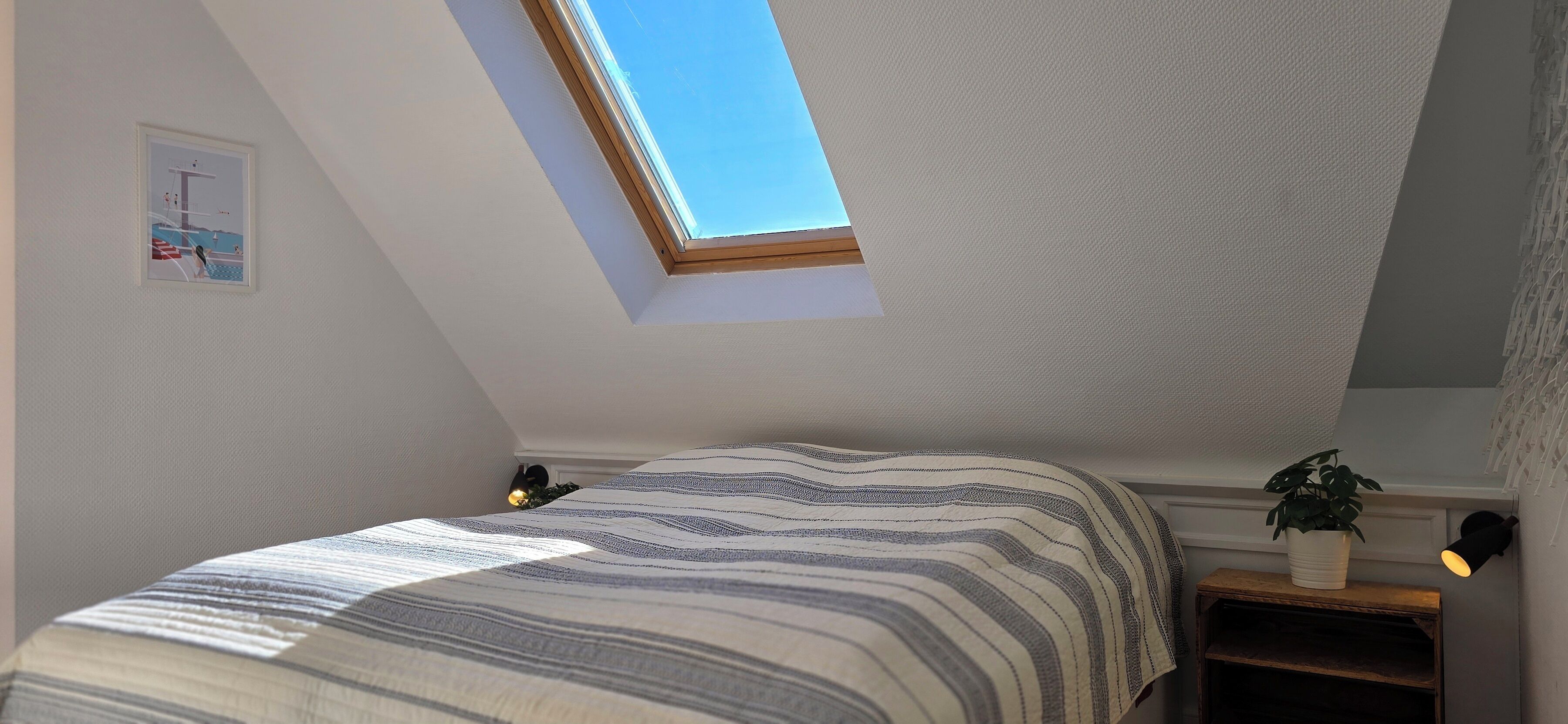 A bedroom with a skylight, a bed with striped bedding, a small bedside table with a plant, and a wall-mounted light.