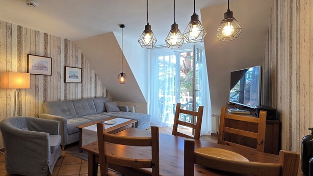 Una sala de estar con sofá, sillones, una mesa de centro y televisor. Se observan listones de madera en las paredes y una ventana con cortinas.