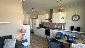 Una sala de estar moderna con un sofá gris, una mesa de comedor con platos azules y una zona de cocina con armarios blancos y un reloj en la pared.