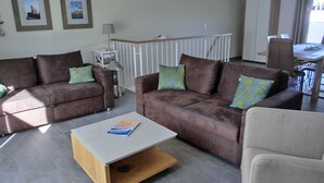 A living room with two brown sofas, a white coffee table, and a dining area with a table and chairs. - 3- Zi.-fewo mit 2 Schlafzimmern auf 2 Etagen für 4 Pers. mit Wlan (Graal-Müritz)