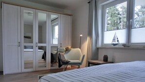 Ein Schlafzimmer mit einem großen Bett, einem Kleiderschrank, einem Sessel und einem Fenster mit Blick auf Bäume.