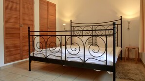 Ein Bett mit Metallgestell und weißer Bettwäsche in einem Zimmer mit Fliesenboden, Holzschränken und einem kleinen Nachttisch.