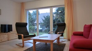 Ein Wohnzimmer mit zwei Sesseln, einem roten Sofa, einem Holztisch und einem Fernseher auf einer Anrichte.