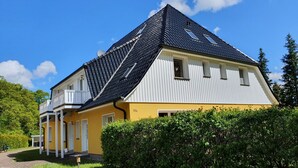 Ein zweistöckiges Haus mit gelber Fassade und schwarzem Dach, umgeben von viel Grün.