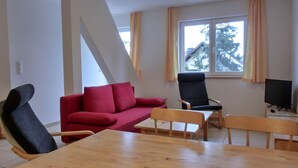 Una sala de estar con un sofá rojo, una silla negra, una mesa de comedor de madera y un televisor sobre un mueble.