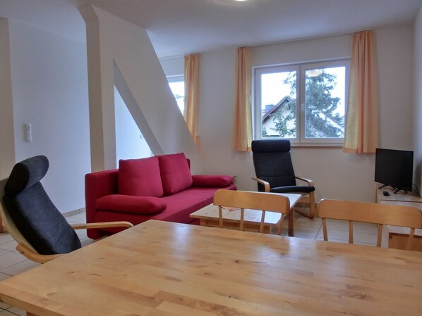 Un salon avec un canapé rouge, un fauteuil noir, une table à manger en bois et un téléviseur posé sur un meuble.