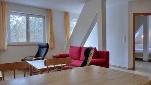 A room with a dining table, two chairs, a red sofa, and a black chair. There is a window with curtains, a door leading to another room, and a white wall with a triangular architectural feature. - 2-zimmer-ferienwohnung für 4 Personen im 1. Obergeschoss -mit Wlan (Neu Hirschburg)