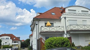 Un immeuble résidentiel de deux étages, avec un toit recouvert de tuiles rouges, des murs extérieurs blancs et un balcon agrémenté d’un parasol jaune.