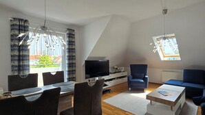 Un salon moderne comprenant un téléviseur, un canapé, une table à manger avec des chaises et une grande fenêtre avec des rideaux.