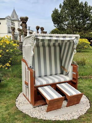 A striped outdoor lounge chair with a wooden frame and a white and gray striped cushion, placed on a gravel path with a garden and a building in the background.
