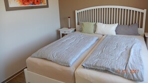 Una cama doble con cabecera blanca y dos almohadas, una mesita de noche con lámpara y un cuadro en la pared.