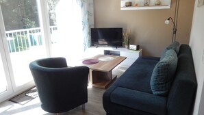 Una sala de estar con un sofá, un sillón, un televisor, una mesita y un ventanal con cortinas.