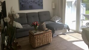 En stue med en grå sofa, et flettet sofabord med en vase med blomster, og et maleri af en strandscene på væggen.