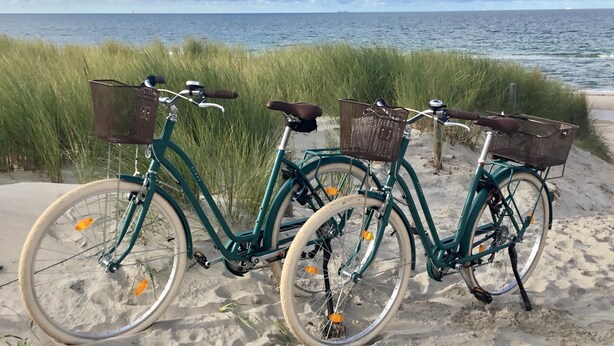 To cykler parkeret på en sandstrand med en græsmark og havet i baggrunden.