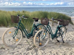 Two bicycles parked on a sandy beach with a grassy area and the ocean in the background.