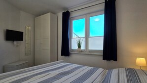 Ein Schlafzimmer mit einem Bett, einem an der Wand montierten Fernseher, einem Kleiderschrank, einem Fenster mit Vorhängen und einer Nachttischlampe.