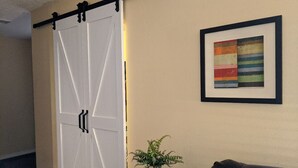 A room with a white sliding door, a framed abstract painting, a potted plant, and a dark-colored sofa. - Spacious 1 Bedroom Condo With Cute Bonus Room (St. George)