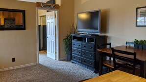 A living room with a TV on a black stand, a wooden dining table, and a door leading to another room. - Spacious 1 Bedroom Condo With Cute Bonus Room (St. George)