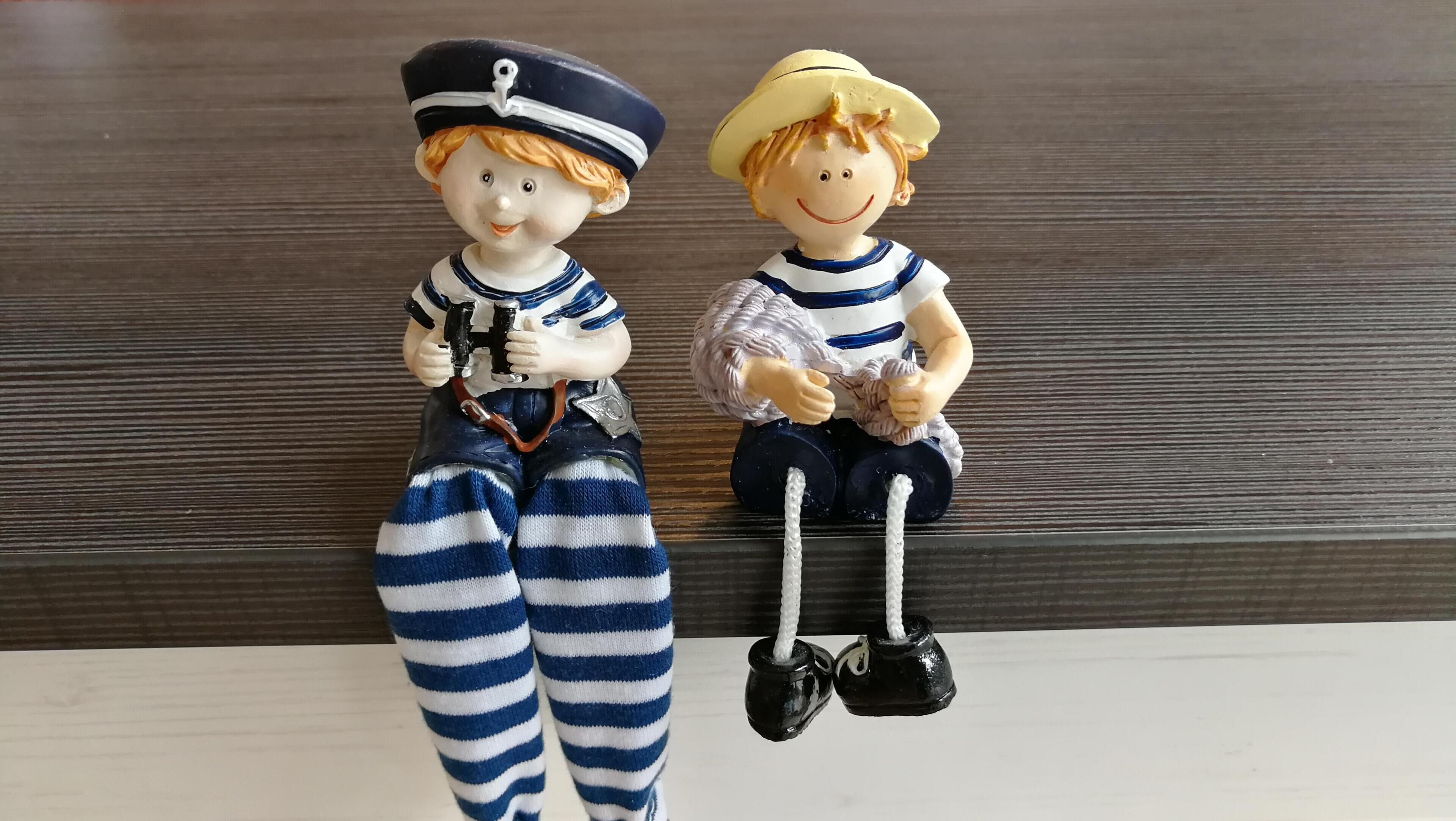 Duas figuras, uma vestida de marinheiro e a outra com roupa listrada e chapéu, sentadas sobre uma superfície de madeira.