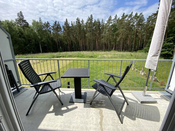 Ein Balkon mit zwei Stühlen und einem Tisch, der auf eine Wiese und einen Wald blickt.