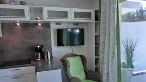 A kitchenette with a flat-screen TV, a wicker chair with a green cushion, and a potted plant by the window. - 2-zimmer-fewo für 2 Pers. im EG mit 2 Fahrrädern (Graal-Müritz)