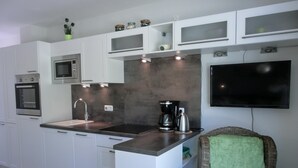 A modern kitchen with white cabinets, a built-in oven, a sink, a coffee maker, and a flat-screen TV mounted on the wall. - 2-zimmer-fewo für 2 Pers. im EG mit 2 Fahrrädern (Graal-Müritz)