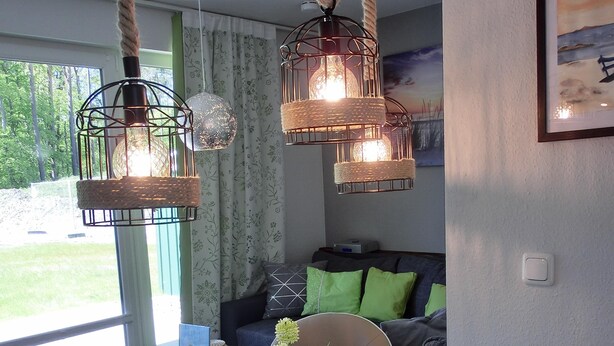 A dining area with a round table set for four, surrounded by chairs. There are hanging light fixtures above the table, and a sofa with cushions in the background. - 2-zimmer-fewo für 2 Pers. im EG mit 2 Fahrrädern (Graal-Müritz)