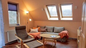 A cozy room with a sofa, two armchairs, a small table, and a floor lamp. There are two windows providing natural light.