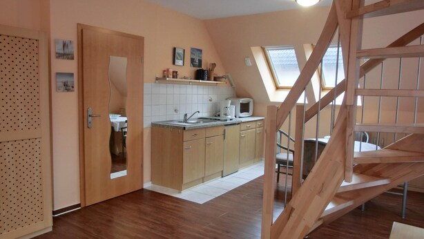 A compact kitchen with wooden cabinets, a sink, and a microwave. There is a small dining area with a table and chairs. A wooden staircase leads to an upper level.