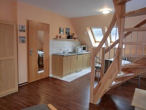 Una cucina compatta con mobili in legno, un lavello e un forno a microonde. C'è una piccola zona pranzo con tavolo e sedie. Una scala in legno conduce a un livello superiore.