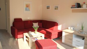 Un salon avec un canapé rouge, une petite table basse et un bureau avec des tiroirs. Les murs sont ornés de tableaux encadrés, et une étagère contient des livres et un vase avec des fleurs.