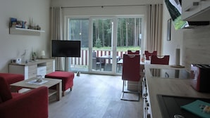 Une chambre d’hôtel moderne dotée d’une grande porte-fenêtre coulissante donnant sur une terrasse extérieure, d’un téléviseur à écran plat, d’un coin repas avec des chaises rouges et d’une kitchenette.