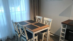 Et spiseområde med et træbord og fire stole, et køkken med komfur, og et vindue med lette gardiner.