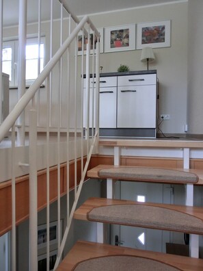 Un escalier avec une main courante en bois et une balustrade blanche. On y trouve un coin cuisine équipé d’un réfrigérateur, d’un évier et d’un plan de travail.